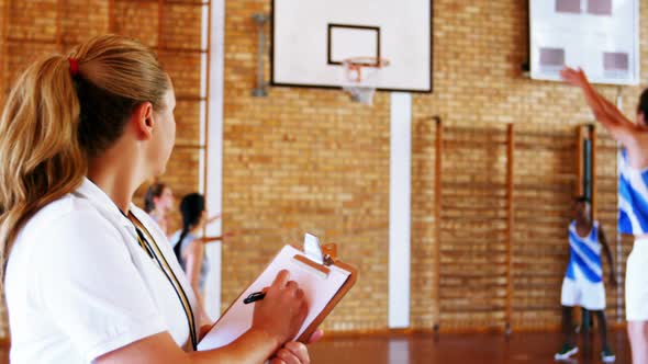 Female coach writing on clipboard while students playing in basketball court