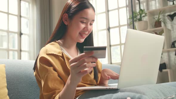 Happy Asian Woman Sitting On Couch Holding Credit Card And Using Laptop Computer Shopping Online
