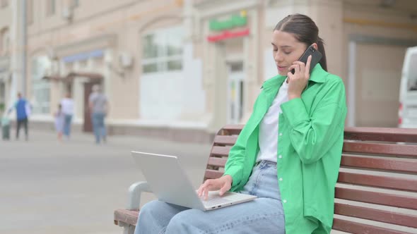 Hispanic Woman Talking on Phone and Using Laptop While Sitting Outdoor on Bench