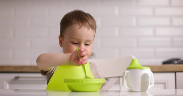 Small Cute Baby Boy Sitting at a Table and Eats Oatmeal with a Spoon