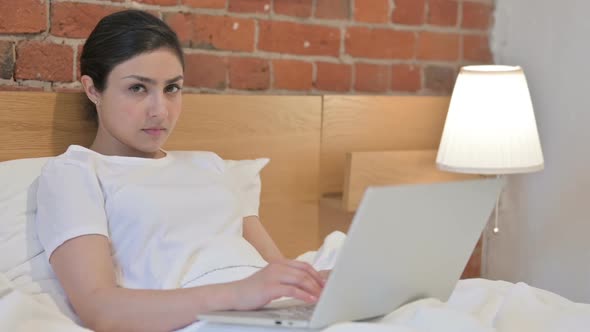 Young Indian Woman with Laptop showing Thumbs Down in Bed