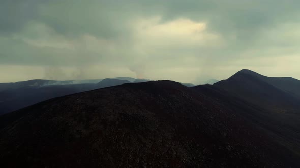 Silhouetted Mountainscape With Distant View Of Erupting Volcano Covered With Smokes. - Aerial Drone