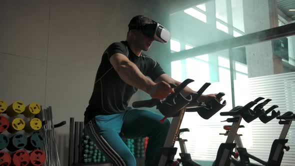 Sporty Male Performing a Heavy Exercise on Bike Simulator in VR Helmet