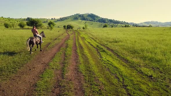 Aerial: Low Angle, Rear View, Flying Forward: Sunburnt Man with Bare Torso, Moves on Horse, Along