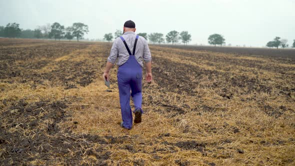 a Man Farmer Walks Through a Plowed Field in the Evening at Sunset Dressed in Blue Overalls Holding