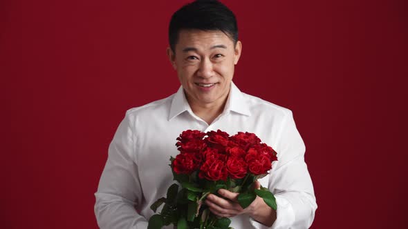 Smiling Asian man in white shirt holds out a bouquet of red roses to the camera