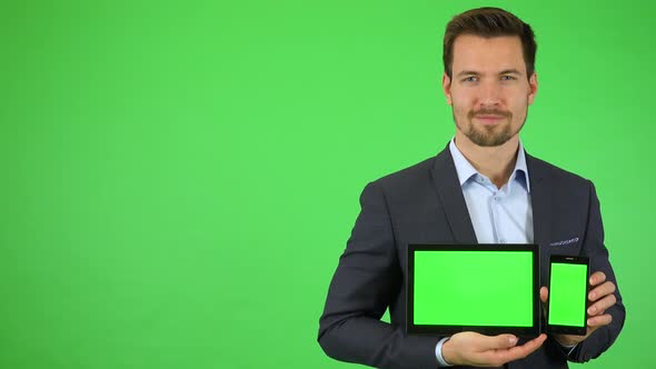 A Businessman Smiles at the Camera and Holds Out a Tablet and a Smartphone with Green Screens