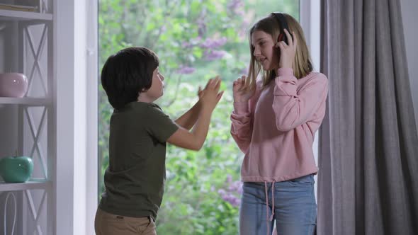 Little Brother Asking Teenage Sister to Share Headphones As Smiling Sibling Dancing Listening to