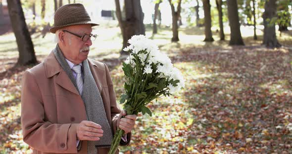 Elegant Senior Man Corrects a Bouquet of Flowers and Talks During Waiting Woman