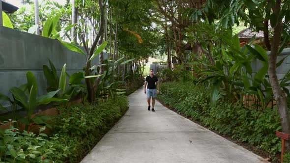 Travel Man in Casual Walking on Path Among Palm Trees in Modern Luxury Resort