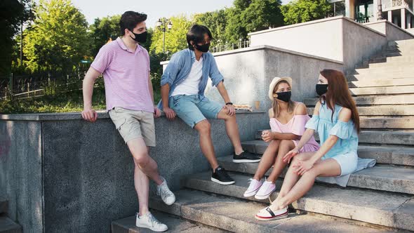 Multiracial Young Friends in Black Protective Masks are Chatting Having Rest on Stairs of Park