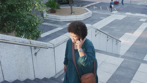 Happy plus size biracial woman walking on stairs and talking on smartphone in city