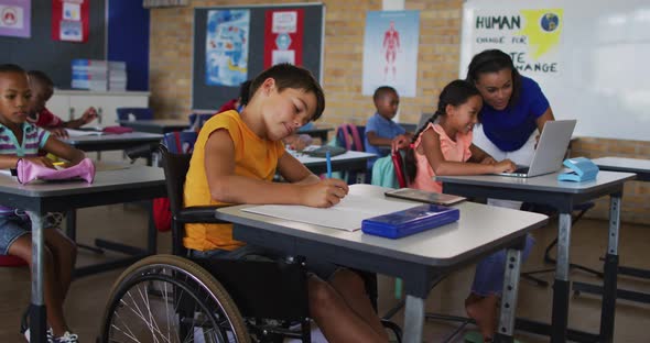 Mixed race schoolboy in wheelchair, sitting in classroom making notes, teacher in background