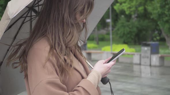 Side View of Concentrated Caucasian Brunette Girl Using Smartphone Outdoors on Rainy Day. Close-up