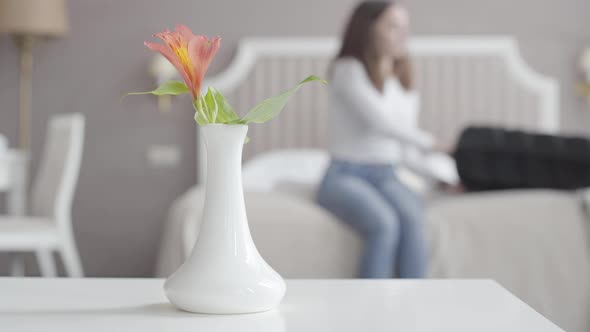 Close-up of Flower in Vase Standing at White Table with Blurred Woman Sitting on Bed and Opening