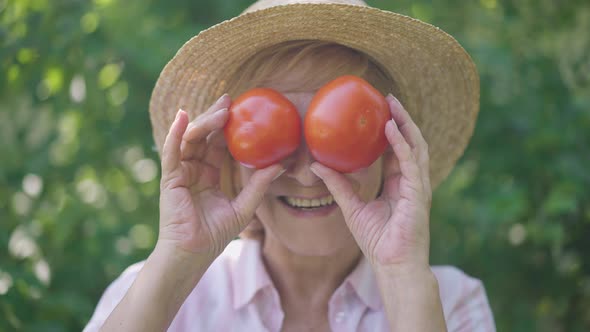 Headshot of Joyful Senior Woman Covering Eyes with Tomatos Having Fun in Garden Outdoors