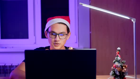 A Young Man in Glasses and a New Year's Hat with a Laptop at the Table. A Student on New Year's Eve