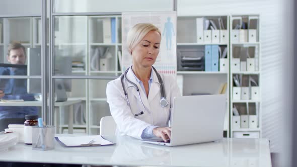 Female Doctor Typing on Laptop and Posing for Camera at Desk