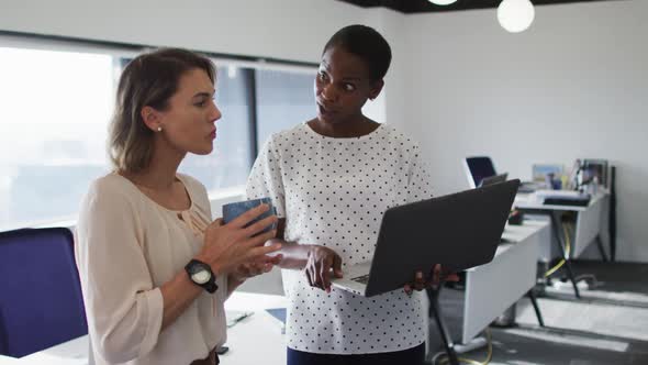 Two diverse female colleagues looking at laptop and discussing in office