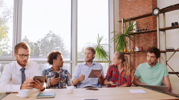 Five Interracial Friends Sitting at Table with Mobile Phones Tablets Laptop Speaking Smiling Shaking