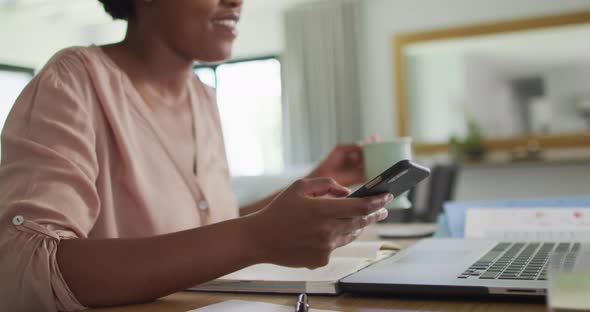 Happy african american woman sitting at table using smartphone and drinking coffee