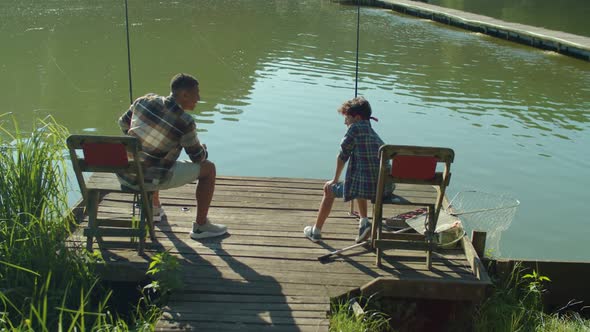 Rear View of Black Dad and Adorable Boy Talking and Fishing on Pond at Dawn