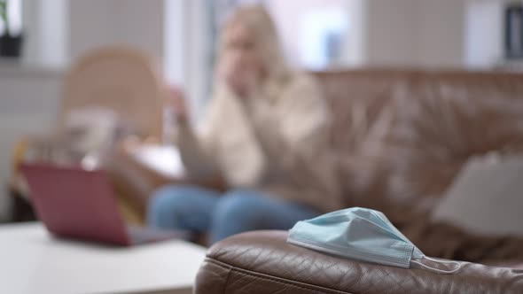 Closeup Coronavirus Face Mask Lying on Couch with Blurred Woman Entering Sitting Down Working Online