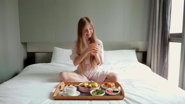 Smiling Woman Sitting on Bed with Two Trays Full of Food Drinking Orange Juice