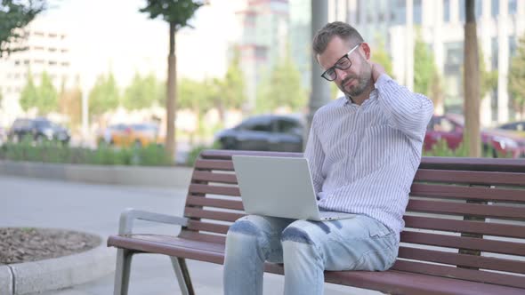 Young Adult Man with Neck Pain Using Laptop While Sitting Outdoor on Bench