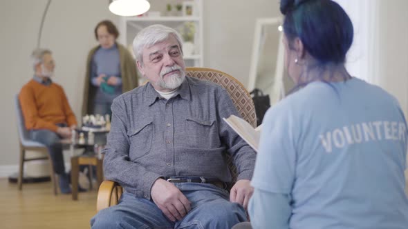 Portrait of Smiling Old Caucasian Man Listening To Volunteer Reading Book. Young Woman Spending Time