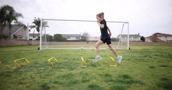 A young girl is warming up for practice by doing high Kees over a set of speed hurdles.