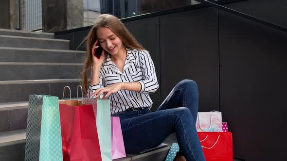 Girl Sitting on Stairs with Bags Talking on Mobile Phone About Sale in Shopping Mall in Black Friday