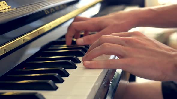 A close up of a man playing a black piano with his hands and fingers in the light of a window.