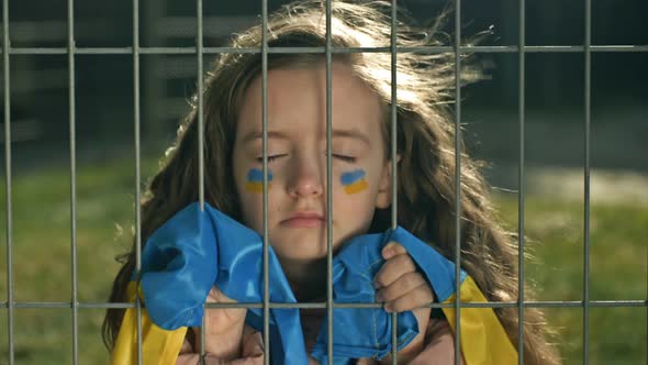 Little Girl is Covered with a Ukrainian Flag with Her Eyes Closed Behind a Metal Grill