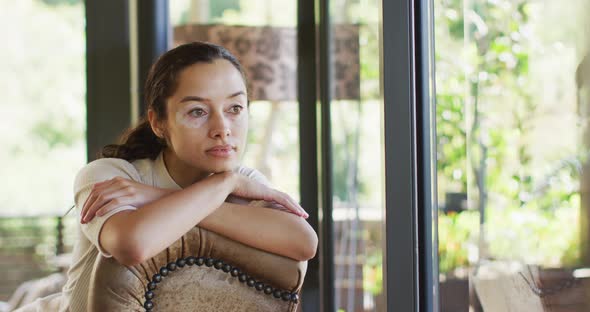 Thoughtful biracial woman with vitiligo sitting on sofa at window and looking outside