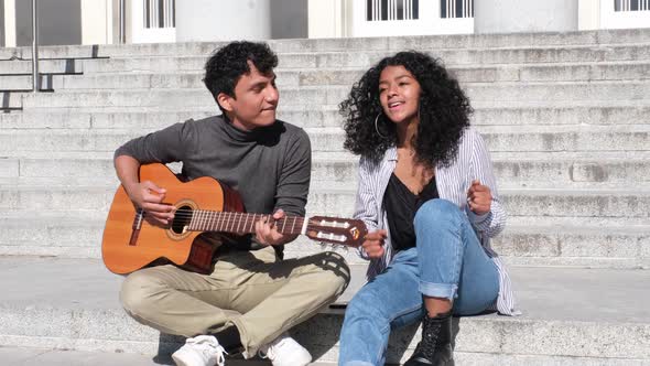 A young latin couple playing the guitar and singing sitting on stairs.