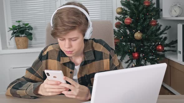 A Teenage Boy with Headphones is Holding a Mobile Phone in His Hands and Studying Information in It
