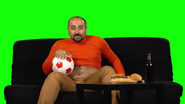 Male Football Fan Watching Soccer While Sitting on Sofa Near Table with Fast Food and Bottle of Beer