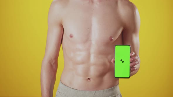 Abs of an Athletic Man on Yellow Background in Studio Raises Phone with Green Screen in Hand at