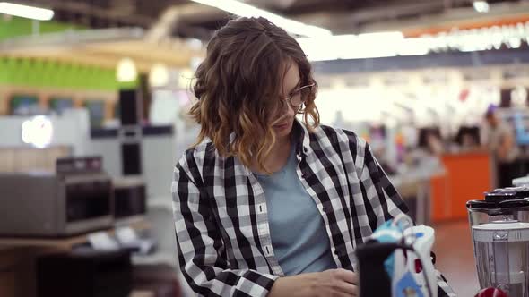 In the Appliances Store a Brunette Curly Woman in a Plaid Shirt Chooses a Blender for Shopping By