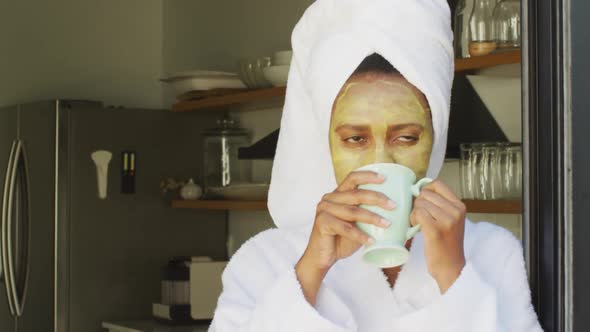 Happy african american woman with beauty mask on face, drinking coffee in kitchen