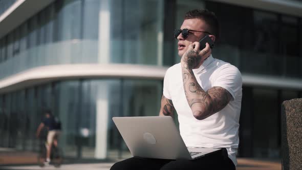 Tattooed Businessman Talking on Phone and Using Laptop for Work, Business Center on Background