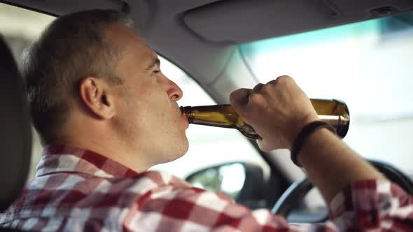 Back Angle View of Depressed Greyhaired Man Drinking Beer From Bottle Sitting on Driver's Seat in