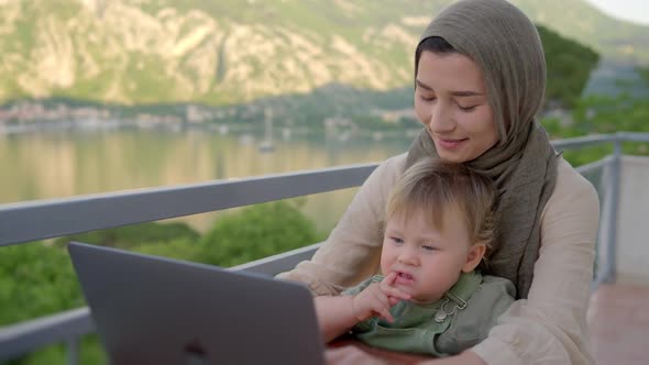 Mother Multitasking Holding Baby Infant and Using Computer Laptop at Home