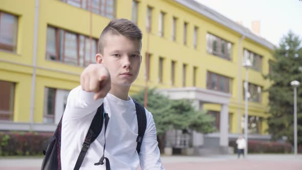 A Caucasian Teenage Boy Points at the Camera and Nods  a School in the Background