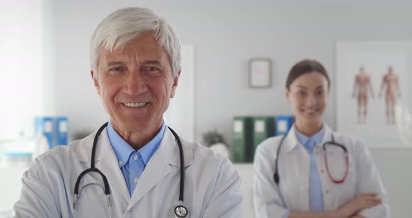 Smiling Senior Man Doctor Looking at Camera with Young Female Colleague Standing on Background