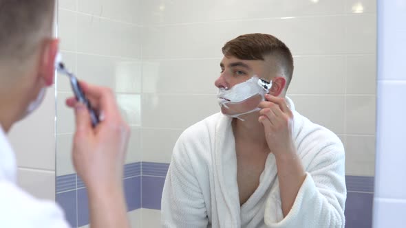 A Young Man Shaves His Face Hair in Front of a Mirror. A Man in a White Coat with Foam on His Face