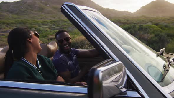 Diverse couple driving on sunny day in convertible car raising their arms in the air and smiling