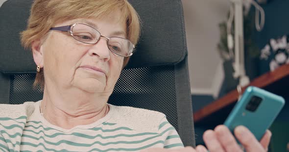 An Elderly Woman Sits in a Chair and Looks Into a Smartphone