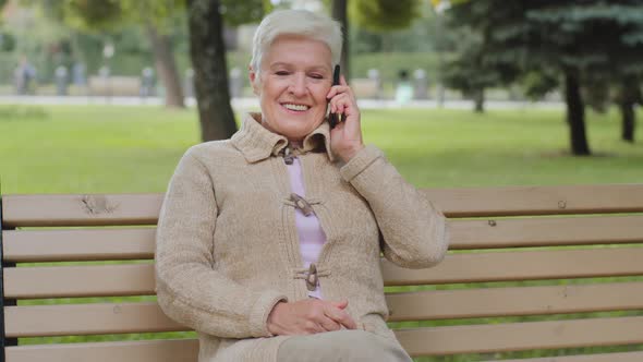 Happy Elderly Woman with Beautiful Face Sitting on Park Bench Takes Phone Call Grayhaired Lady of